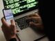 unrecognizable hacker with smartphone typing on laptop at desk